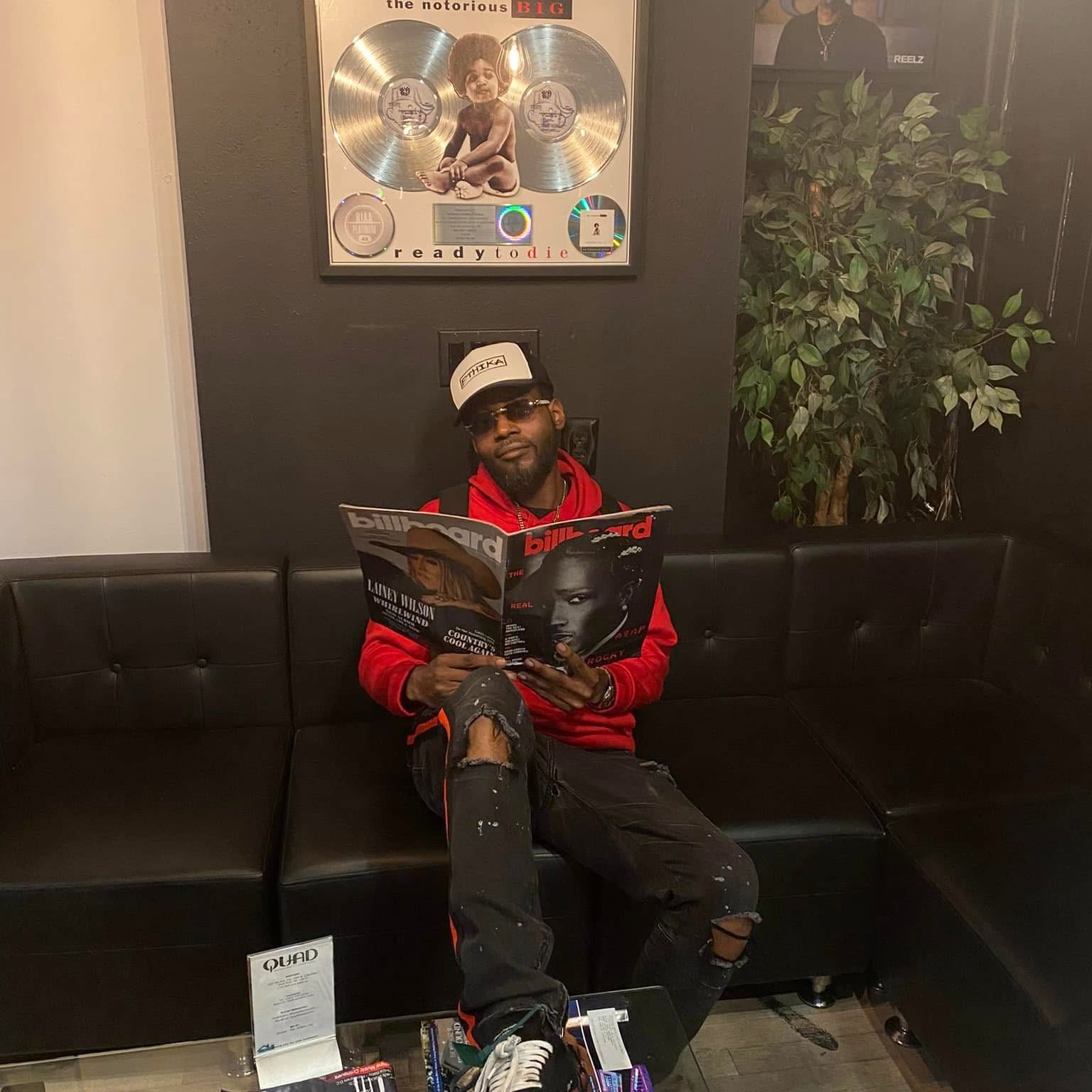 Flyboi Rich seated in a red hoodie holding a magazine